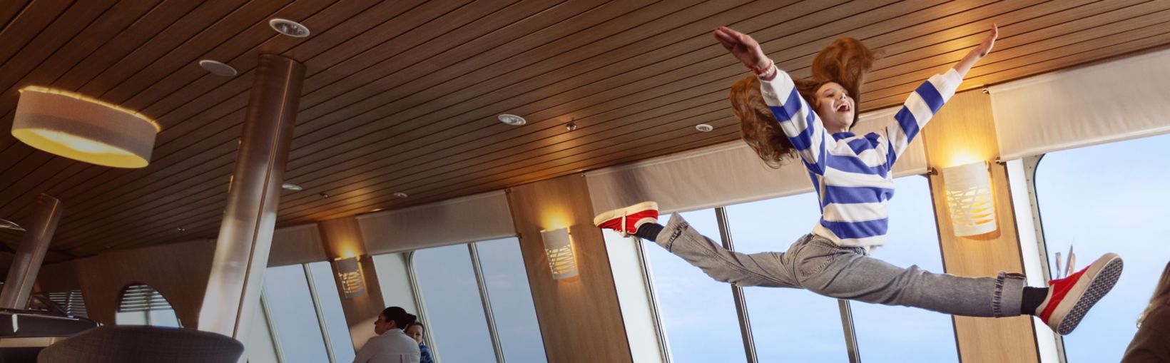 A young girl, with long red hair and wearing a stripy jumper, jumps in the air in the Stena Plus lounge.